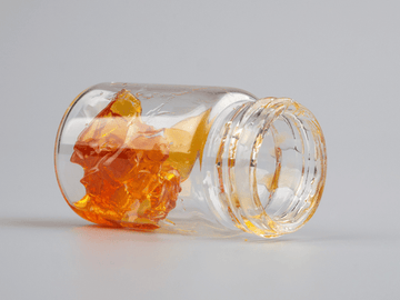 A small clear glass jar tipped over, containing amber-colored cannabis concentrate or THCA extract, displayed on a clean, white background.