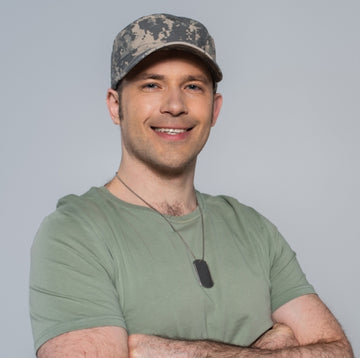 Smiling military veteran wearing a cap and dog tags, standing confidently in a casual pose against a neutral background.