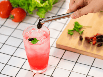 A vibrant pink hemp-infused mocktail garnished with a mint leaf, placed on a white tiled countertop with fresh ingredients like tomatoes and herbs in the background.